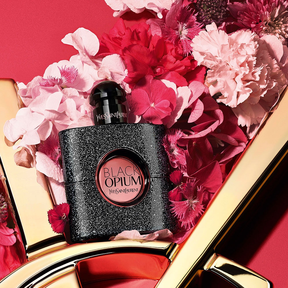 Botella de perfume negra con purpurina Black Opium Yves Saint Laurent rodeada de flores rosas y rojas, sobre un fondo rojo con el detalle del logo dorado de YSL.