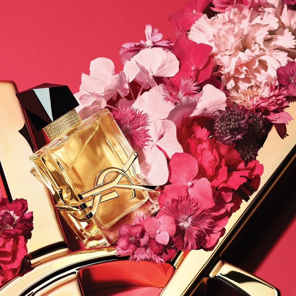 Frasco de YSL Libre Eau de Parfum con logotipo YSL dorado, decorado con flores rosas y rojas sobre un fondo rojo con un detalle del logotipo YSL dorado.