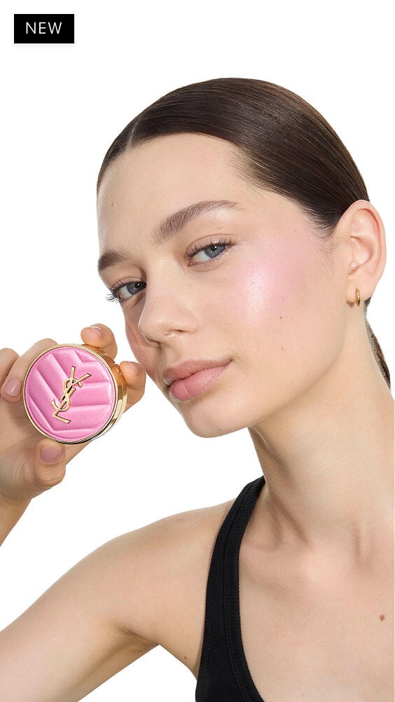 Oksana, de piel clara, sosteniendo un colorete en polvo rosa YSL Make Me Blush en el tono 10, mostrando un rubor sutil en su mejilla.
