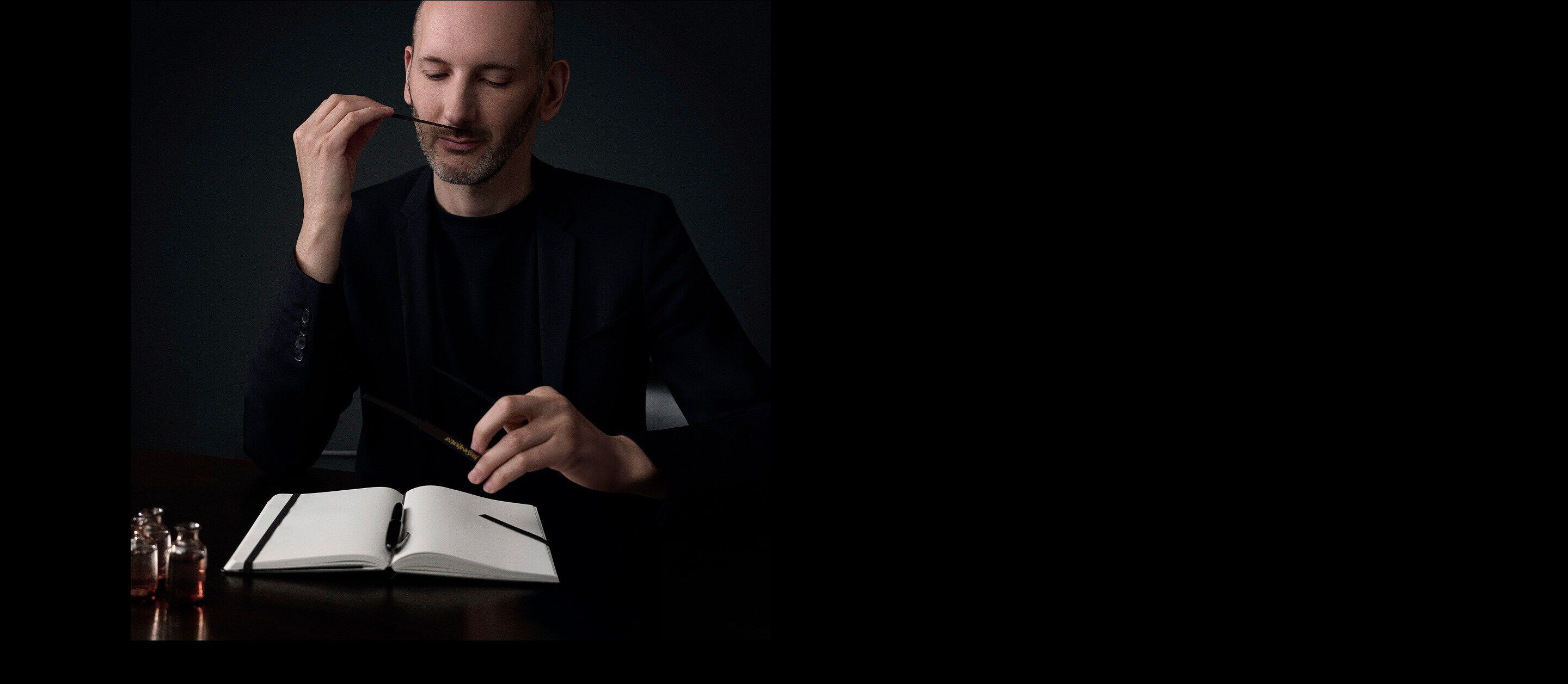 Quentin Bisch, perfumer, in a black outfit smelling a fragrance blotter at a desk with an open notebook and small perfume bottles in a dark setting.