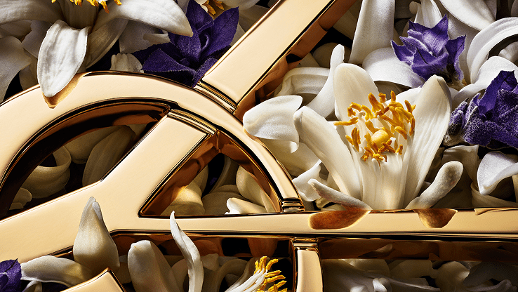Close-up of the gold YSL Cassandre logo nestled among delicate white orange blossoms and vibrant purple lavender flowers.