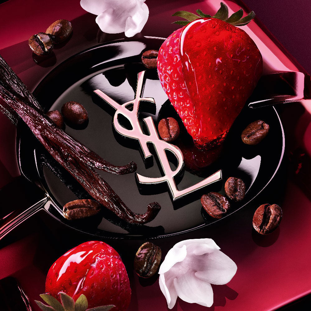 Close-up of fresh strawberries, vanilla pods, jasmine flowers, and roasted coffee beans arranged over the Black Opium Eau de Parfum Pink Glaze bottle with the iconic YSL cassandre.